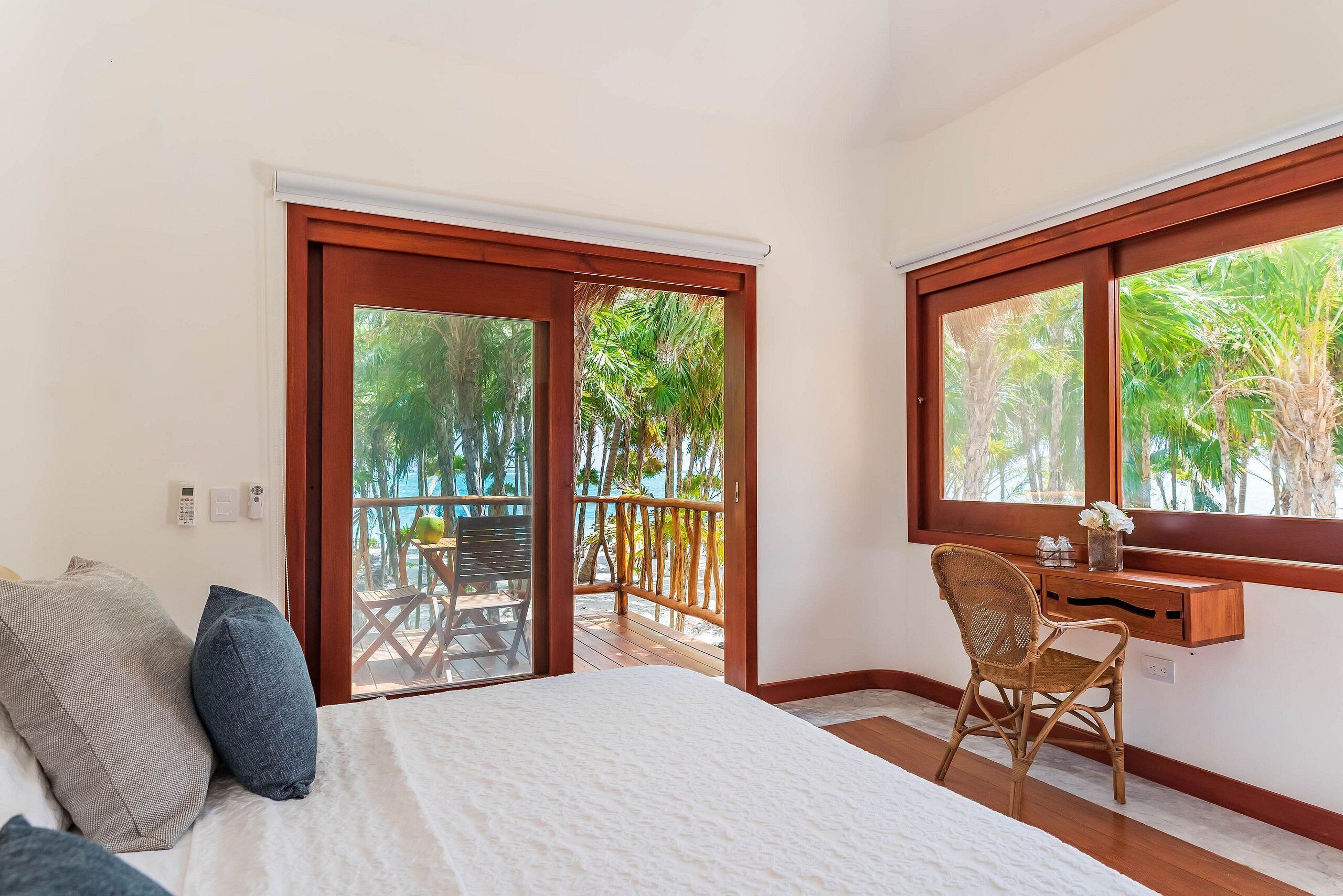 A bedroom with a bed, a desk, and a chair, a balcony with a wooden railing, and a view of palm trees.