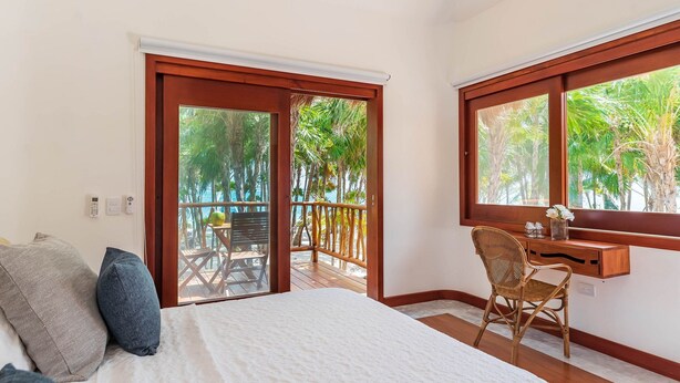 Un dormitorio con cama, escritorio y silla, un balcón con barandilla de madera y vista a palmeras.