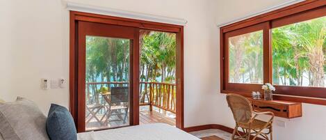 A bedroom with a bed, a desk, and a chair, a balcony with a wooden railing, and a view of palm trees.