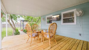 A covered porch with a wooden deck, wicker chairs, a glass table, and a wall-mounted fan.