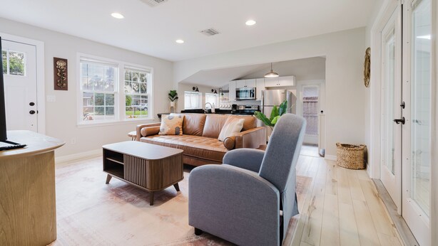 A spacious living room with a sofa, armchair, and coffee table, a kitchen in the background, and large windows providing natural light.