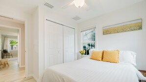 A bedroom with a large bed, white walls, a ceiling fan, and a window with a view of greenery outside.