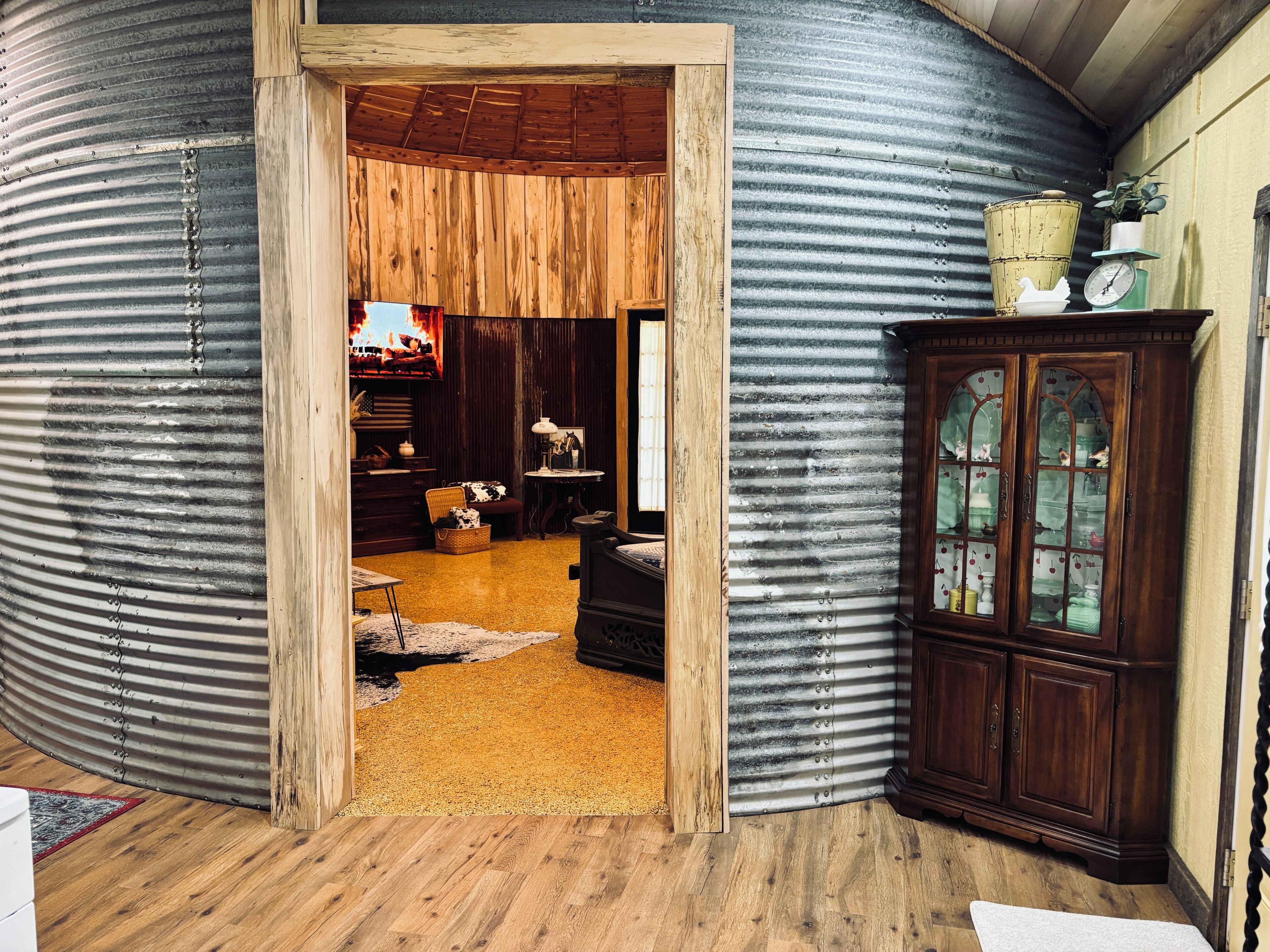 Un intérieur rustique avec une porte en bois donnant sur un espace de vie agrémenté d’une cheminée, d’un meuble en bois aux portes vitrées et d’un téléviseur diffusant une scène de feu.