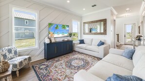 Een woonkamer met een grote televisie, een bank, een kamerkleed met een patroon en uitzicht op een huis.