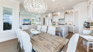 Een ruime eethoek met een houten tafel en beklede witte stoelen, een keuken met witte kasten en roestvrijstalen apparatuur, en een grote kroonluchter.