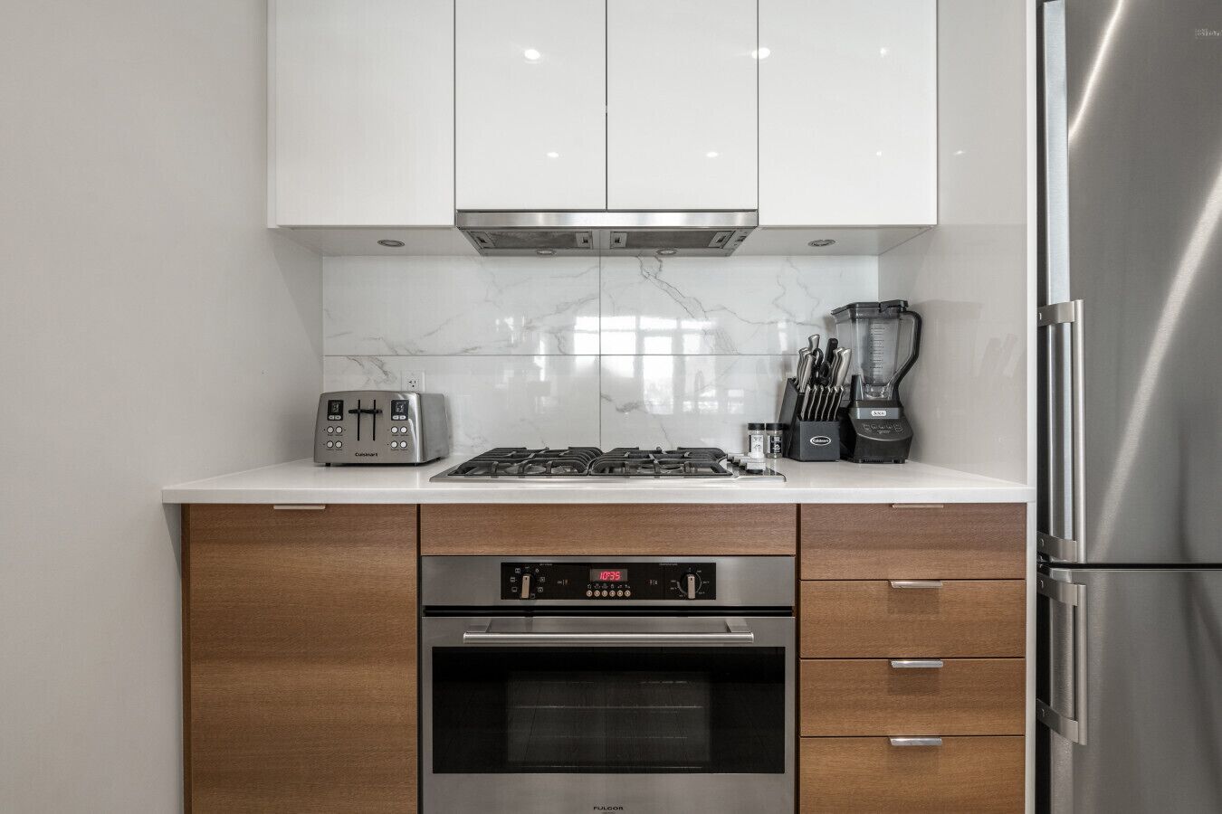 A modern kitchen with white cabinets, a built-in oven, a stovetop, a toaster, and a refrigerator.