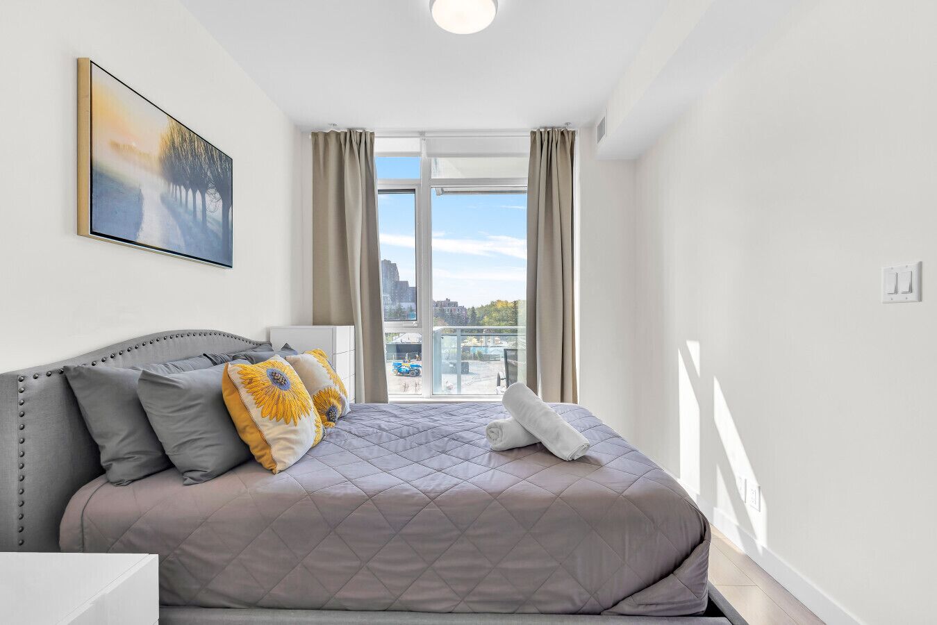 A modern bedroom with a large bed, a city view through the window, and a painting on the wall.