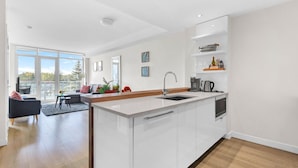 Una cocina moderna con una isla grande, un rincón de comedor con sofá y mesa, y vistas a árboles a través de amplios ventanales.