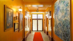 Um corredor de hotel com obras emolduradas nas paredes, tapete vermelho e uma grande pintura abstrata à direita.