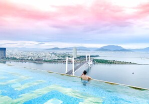 A man relaxing in a large outdoor swimming pool with a cityscape and bridge in the background.