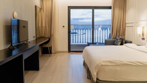 Ett hotellrum med en säng, ett skrivbord med en tv, en balkong med ett räcke och utsikt över himlen.