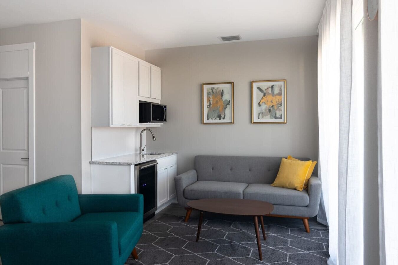 A compact living space with a kitchenette, a teal sofa, a grey sofa with a yellow pillow, and two abstract paintings on the wall.