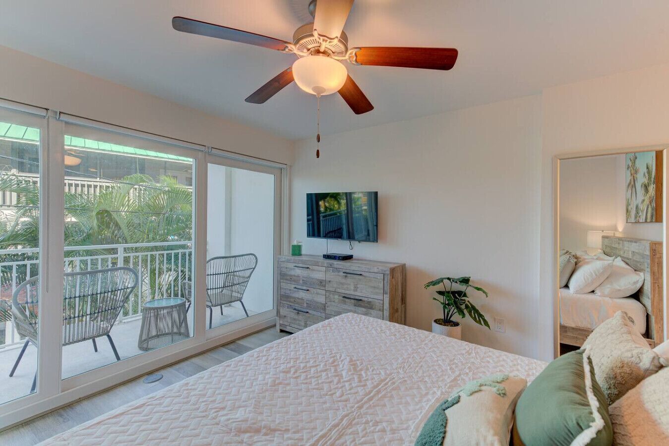 A bedroom with a bed, a ceiling fan, a TV, a dresser, and a balcony with chairs.