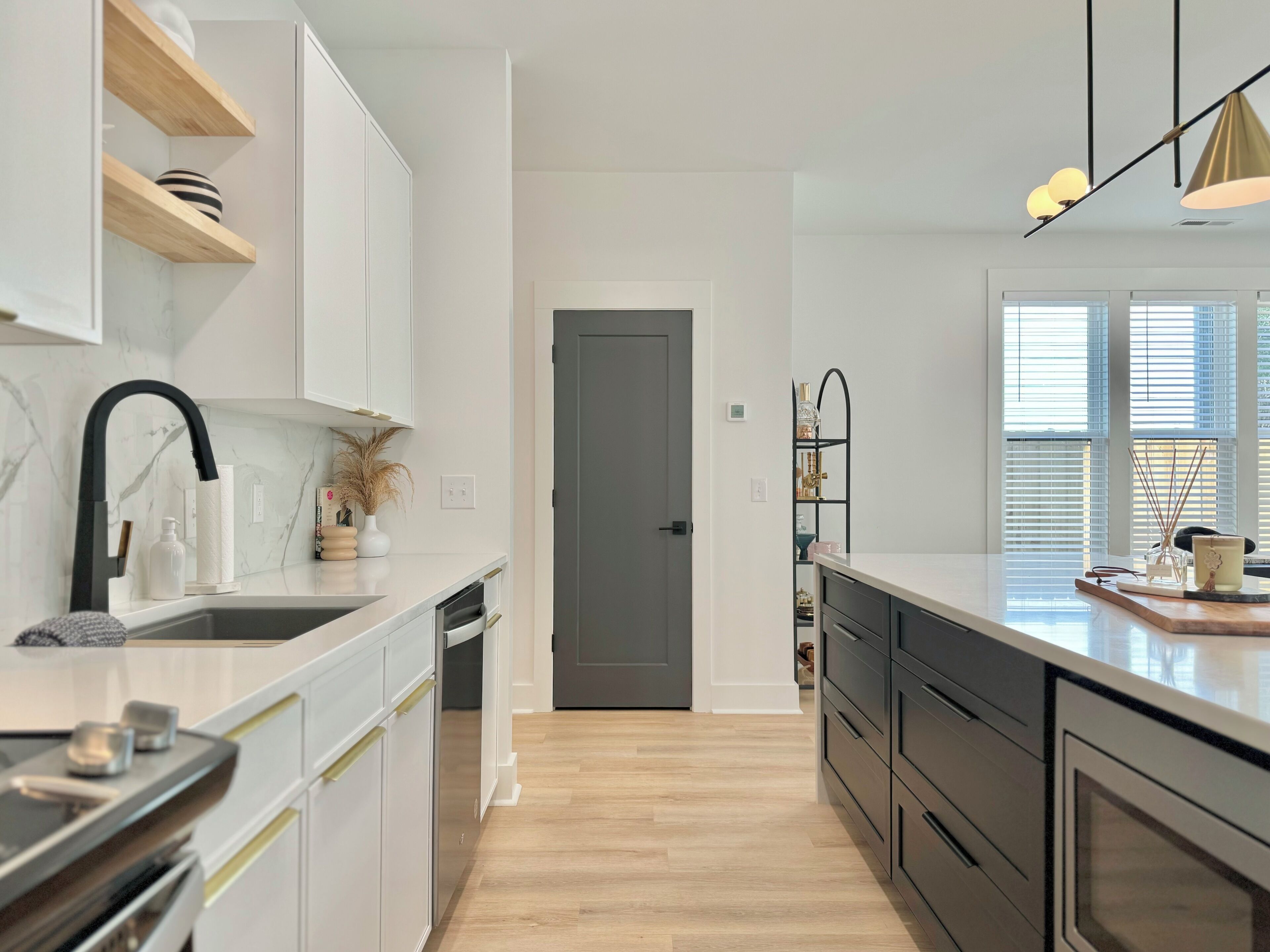 A modern kitchen with white cabinetry, a black faucet, and a wooden floor. There is a door leading to another room and a window with blinds.