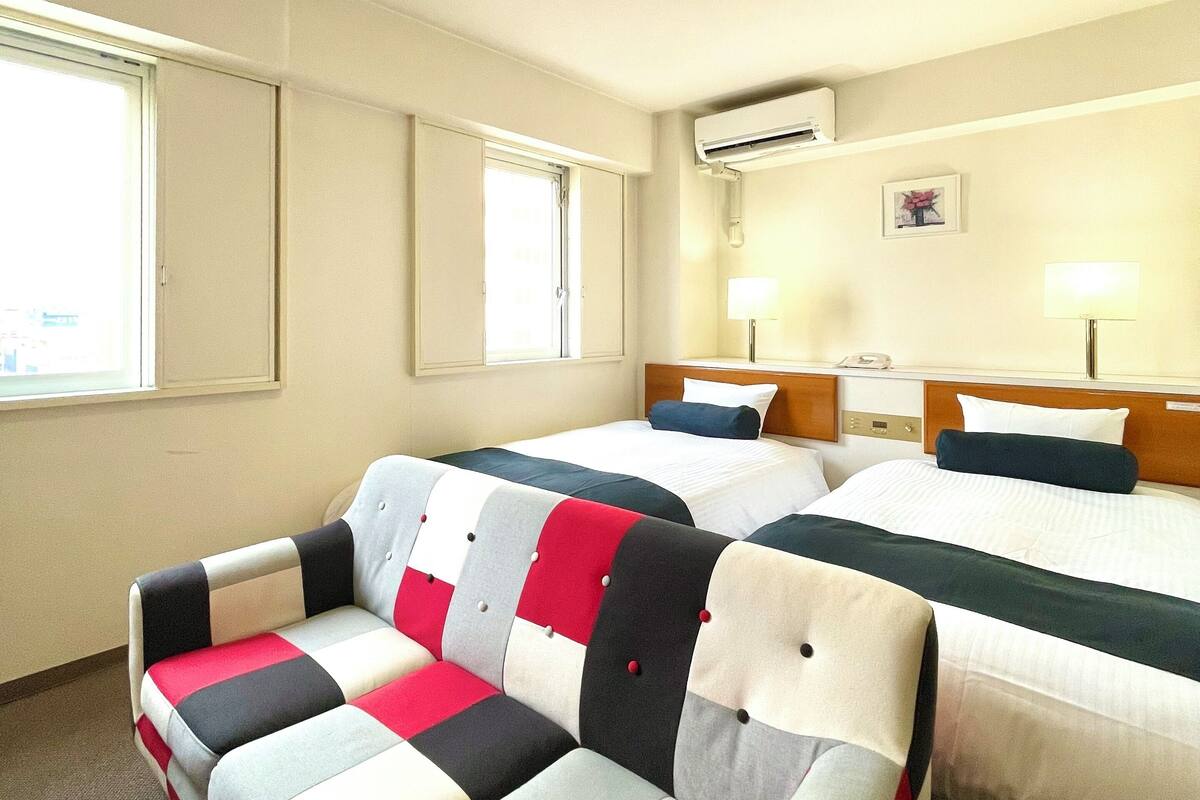 Ein Hotelzimmer mit zwei Betten, einem Sofa, einem Fenster, einer Klimaanlage und einer Lampe.