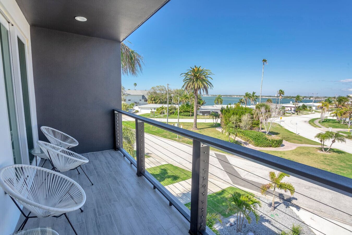A balcony with three white chairs, a grey wall, and a view of a road with palm trees and a body of water.