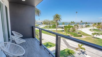 A balcony with three white chairs, a grey wall, and a view of a road with palm trees and a body of water.