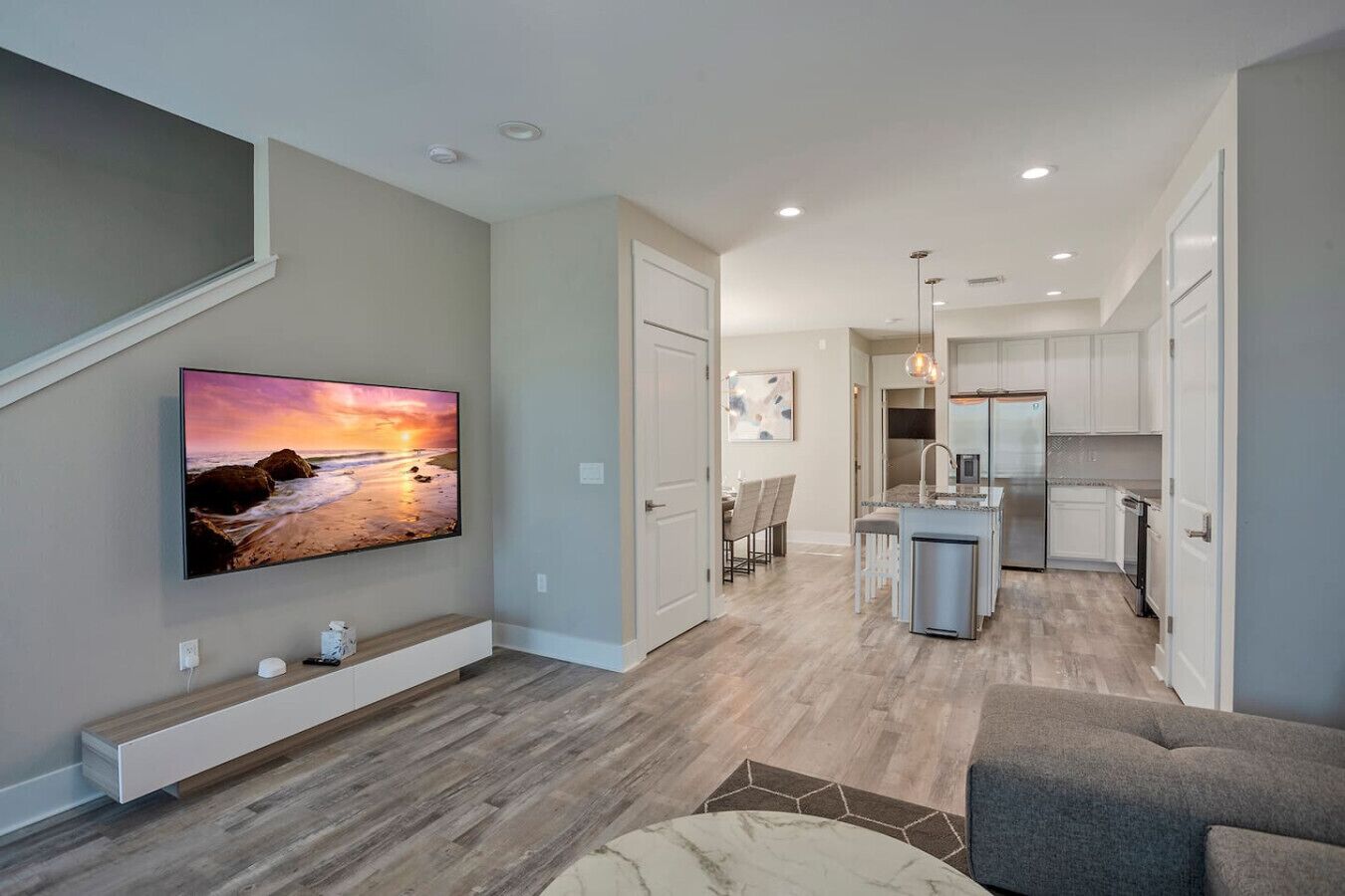 A modern living room with a flat-screen TV, wooden flooring, a gray sofa, and a kitchen with white cabinets and stainless steel appliances.