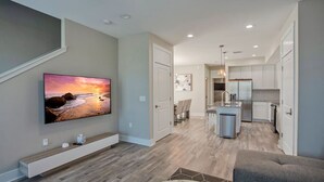 Een moderne woonkamer met een flatscreen televisie, houten vloer, een grijze bank en een keuken met witte kasten en roestvrijstalen apparatuur.