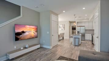 A modern living room with a flat-screen TV, wooden flooring, a gray sofa, and a kitchen with white cabinets and stainless steel appliances.