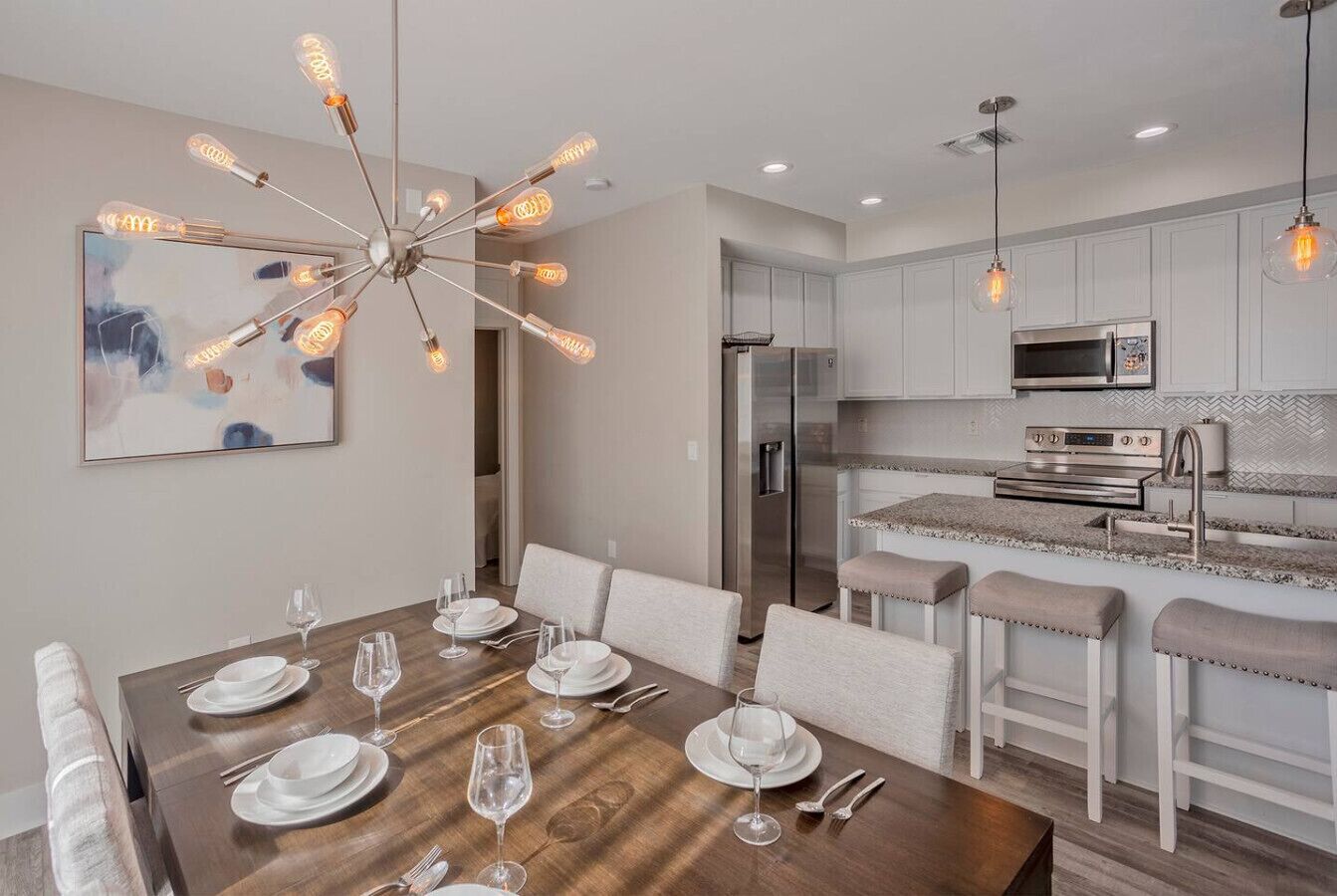 A modern dining area with a wooden table set for six, a contemporary chandelier, and a kitchen with stainless steel appliances.