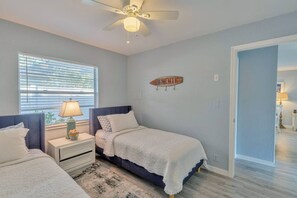 A bedroom with two beds, a ceiling fan, a window with blinds, a bedside table with a lamp, and a wall sign that says "BEACH".