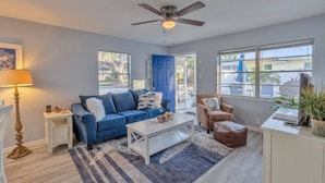 Ein Wohnzimmer mit einem blauen Sofa, einem weißen Couchtisch, einem braunen Sessel, einem Deckenventilator und Blick ins Freie durch große Fenster.