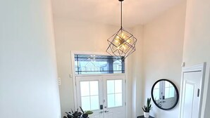 Un couloir avec une porte blanche, un banc, une plante en pot, une console noire, un miroir rond et une suspension lumineuse aux motifs géométriques.