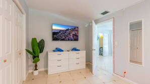 A hallway with a white door, a white dresser with blue towels, a potted plant, and a TV displaying a cityscape. - Tropical Paradise-4/4 (Largo)