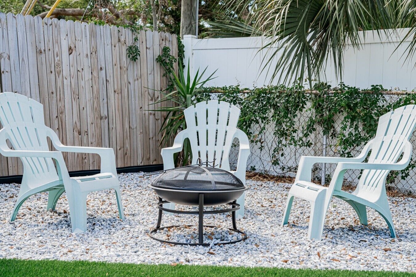 Eine Terrasse mit drei weißen Adirondack-Stühlen, einem runden Tisch und einer Feuerstelle.