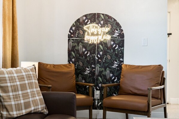 A room with a brown leather sofa, a plaid pillow, and a wall with a neon sign that reads 'This is our happy place.'