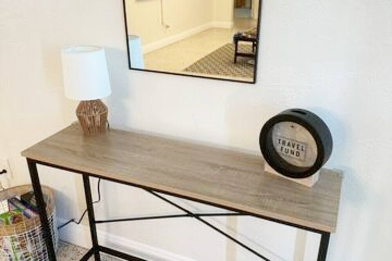 A wooden console table with a lamp and a 'TRAVEL FUND' sign, a mirror on the wall, and a trash bin to the left.