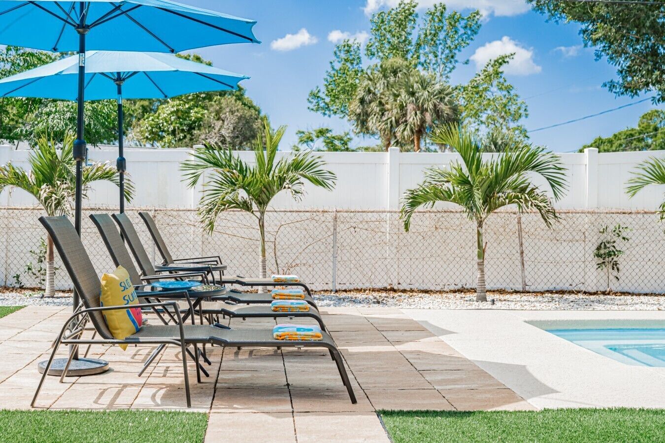 A poolside area with lounge chairs and a blue umbrella, surrounded by a white fence and palm trees.