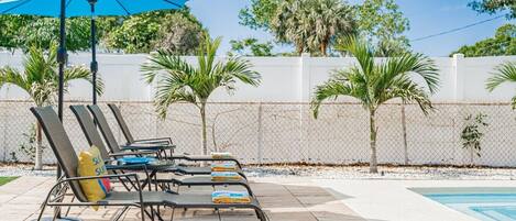A poolside area with lounge chairs and a blue umbrella, surrounded by a white fence and palm trees.