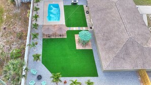 A backyard with a swimming pool, artificial grass, a patio area with tables and umbrellas, and a small tree. - Palm Oasis 2/2 Serene Backyard Private Poolside (St. Petersburg)