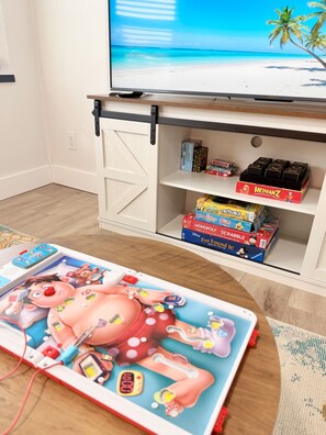 Een kamer met een televisie waarop een strandscène te zien is, een plank met gezelschapsspellen en een tafel met een kinderspeelgoed.