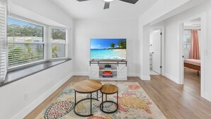 A room with a TV on a white stand, a ceiling fan, a window with blinds, a colorful rug, and a bed in the background. - Chic Coastal Getaway (Largo)