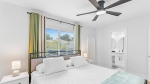 A modern bedroom with a bed, bedside tables, a ceiling fan, a window with curtains, and a bathroom with a mirror and sink. - Chic Coastal Getaway (Largo)