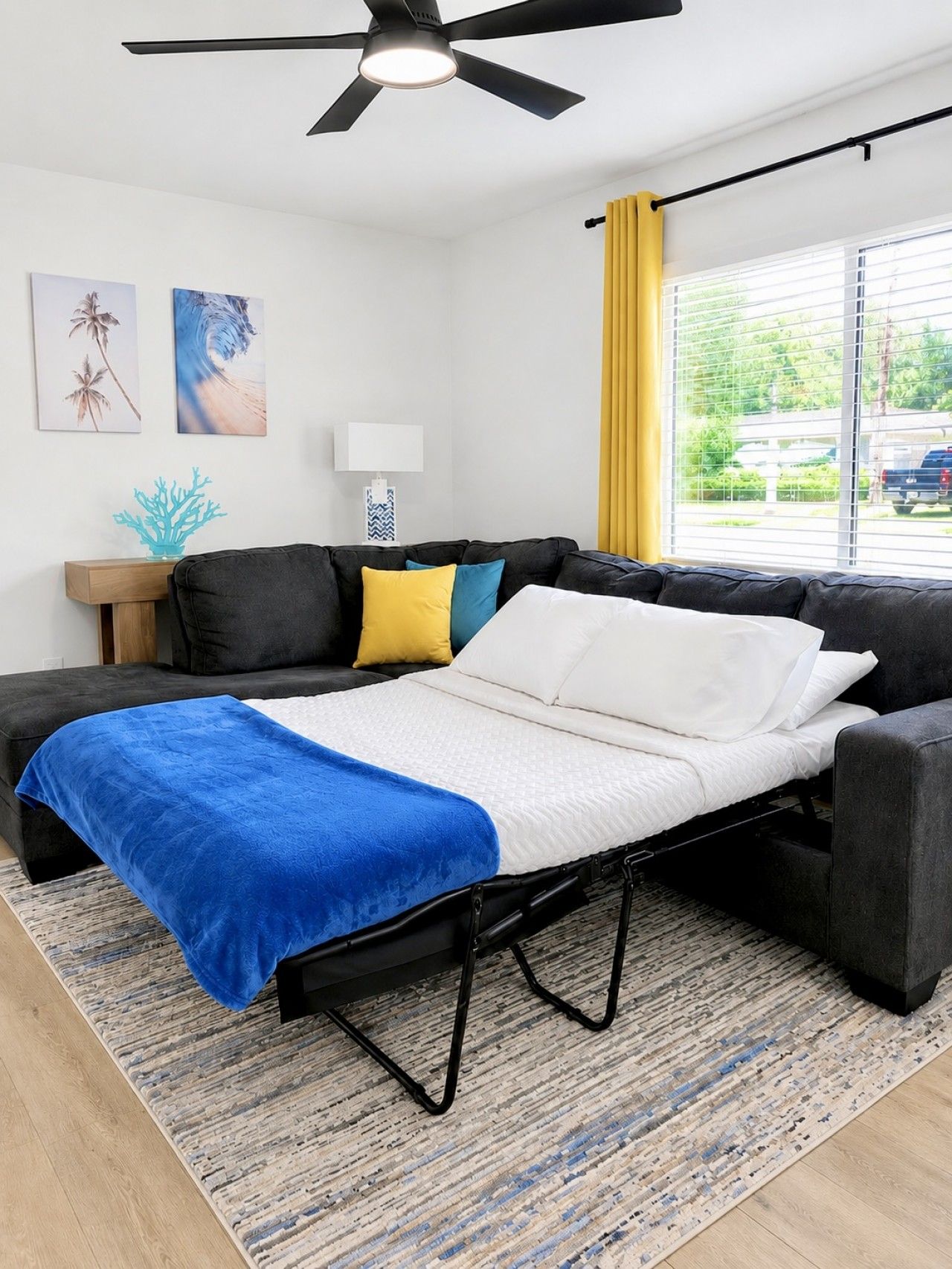 A modern room with a sofa bed, a ceiling fan, artwork on the wall, and a view through a window.