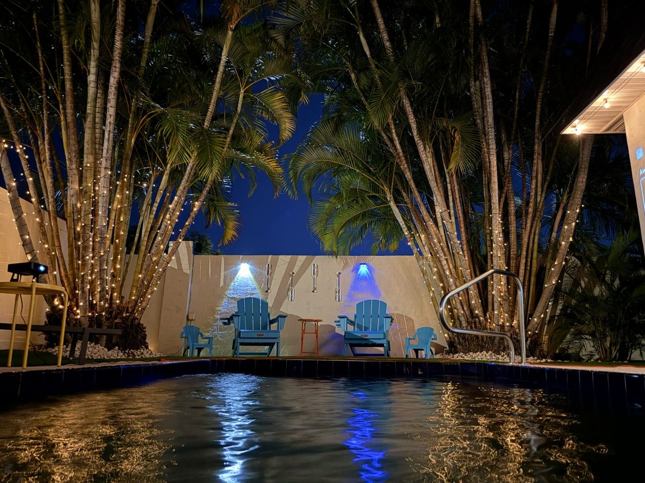 Zona de piscina al aire libre con sillas y una fuente de agua, rodeada de palmeras y luces colgantes.