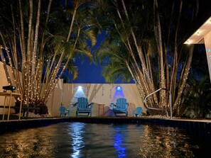 Un espace extérieur avec piscine, des chaises et un élément décoratif aquatique, le tout entouré de palmiers et illuminé de guirlandes lumineuses.