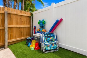 Un jardin avec une pelouse artificielle, une clôture en bois et divers équipements de jeux pour enfants, notamment une glacière Coleman, une piscine gonflable bleue et verte, et un flotteur de piscine rouge et bleu.