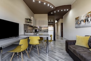 Una sala moderna con una televisión de pantalla plana, un sofá con cojines amarillos y una zona de comedor con sillas amarillas, con una cocina al fondo.