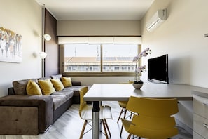 Una sala de estar moderna con sofá, mesa de comedor, sillas, televisor y ventana con vista a edificios.