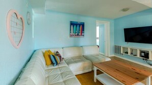 A living room with a white sectional sofa, a wooden coffee table, a flat-screen TV, and a surfboard painting on the wall.