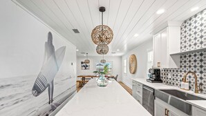 A modern kitchen with a long island, white cabinetry, and a backsplash with a star pattern. There is a large painting of a surfer on the wall, a hanging light fixture, and a plant on the island. - NEW - Ocean View, Heated Pool, Beach, Sleeps 18, Fully Loaded (Gulf Shores)
