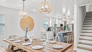 A dining area with a wooden table set for six, a kitchen in the background, and a staircase to the right. - NEW - Ocean View, Heated Pool, Beach, Sleeps 18, Fully Loaded (Gulf Shores)