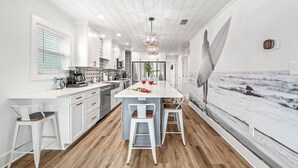 A modern kitchen with white cabinetry, a central island with bar stools, and a large window with a view of the ocean. - NEW - Ocean View, Heated Pool, Beach, Sleeps 18, Fully Loaded (Gulf Shores)