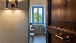 A hallway leading to a kitchen with a window, a door with a brass handle, and a wall-mounted light fixture. - GuestHost - Elegant Apartment Testaccio Roma (Rome)