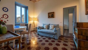 A room with a blue sofa, a desk, a chair, a lamp, and a dining table with a vase of flowers. - GuestHost - Elegant Apartment Testaccio Roma (Rome)